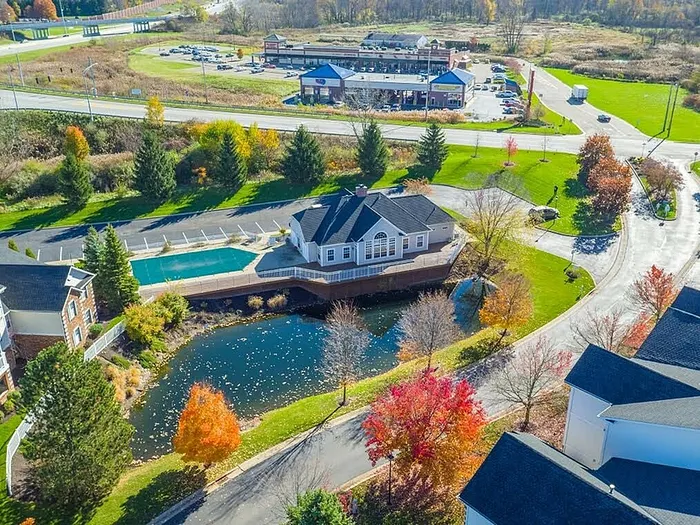 Charming aerial view of a picturesque community featuring a central clubhouse, sparkling pool, and serene pond amid vibrant autumn foliage.