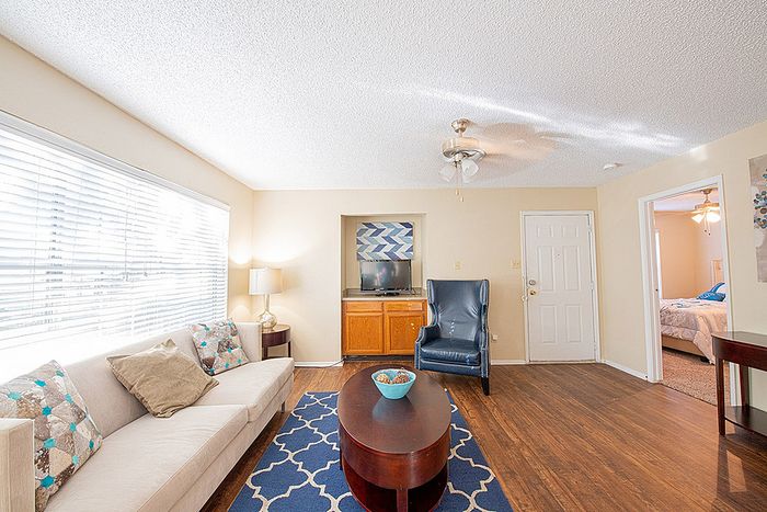Bright living room with elegant hardwood floors, cozy beige couch, and modern decor. Features a stylish blue rug, natural lighting from large windows, and a view into a welcoming bedroom.