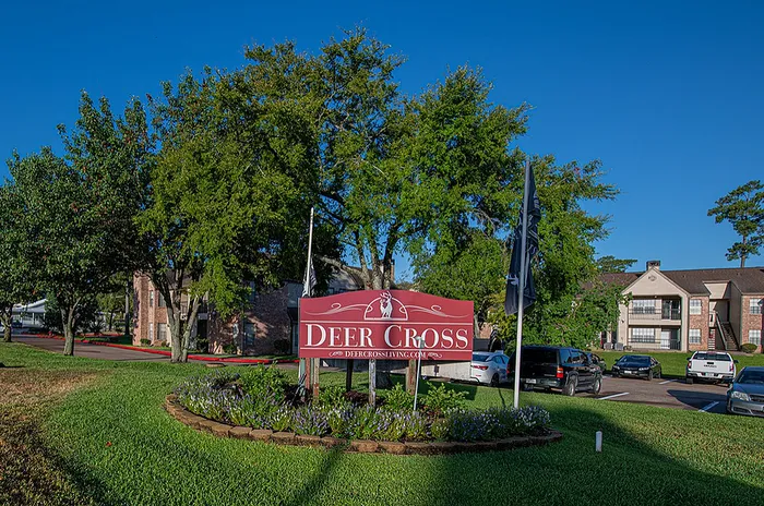 Inviting entrance to Deer Cross Apartments with lush green landscaping and modern residential buildings under a clear blue sky.