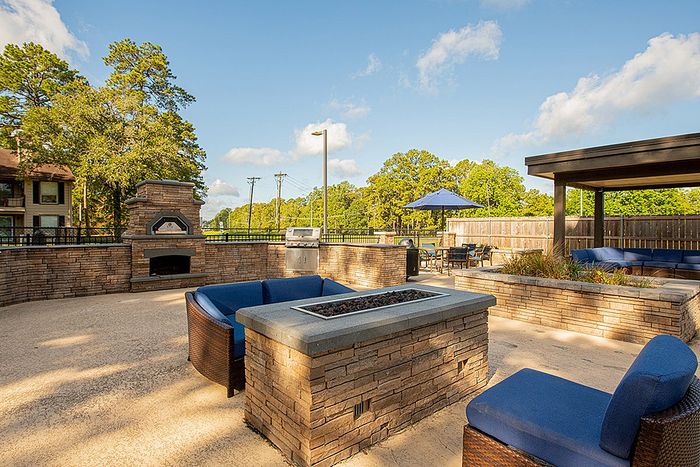 Inviting outdoor patio with cozy blue seating, stone fireplace, and grill area set against a vibrant, green backdrop.