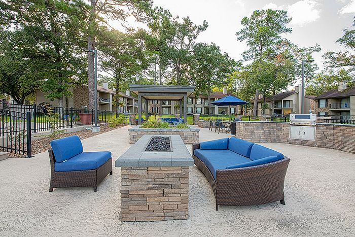Inviting outdoor lounge area with blue cushioned seating, stylish fire pit, and barbecue grill, surrounded by lush greenery and modern apartment buildings.