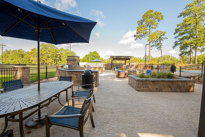 Inviting outdoor patio area with stylish seating, large umbrella, and modern grill, surrounded by lush trees under a clear blue sky.