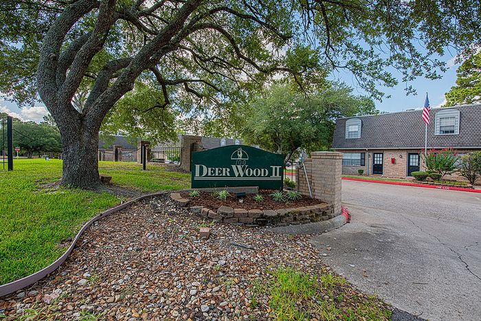 Charming entrance to Deer Wood II community featuring a welcoming sign under a picturesque tree, surrounded by lush greenery and serene pathways.