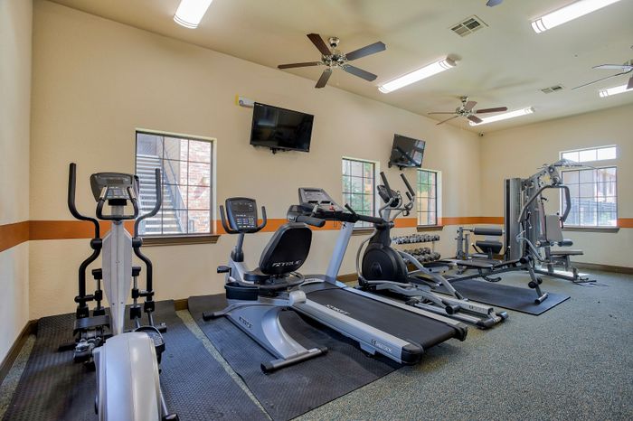 Modern fitness center with state-of-the-art exercise equipment, including treadmills, elliptical machines, and a weight station, set in a bright room with ceiling fans and wall-mounted TVs.