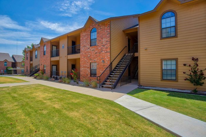 Charming apartment complex with rustic brick exterior, lush green lawns, and bright blue skies, offering a welcoming community vibe.