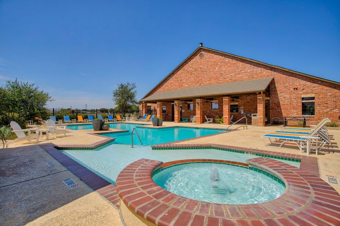 Inviting community pool area with a sparkling jacuzzi and vibrant lounge chairs, set against a charming brick clubhouse backdrop on a sunny day.