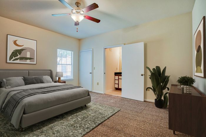 Inviting bedroom with a modern gray bed, lush carpet, chic ceiling fan, and decorative plants, complemented by wall art and natural light from a large window.