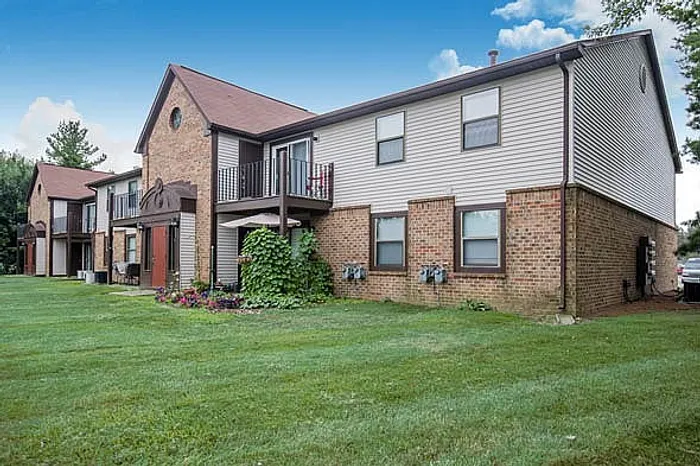 Charming multi-unit brick townhouse with lush green lawn, featuring upper balcony and vibrant flower beds under a clear blue sky.