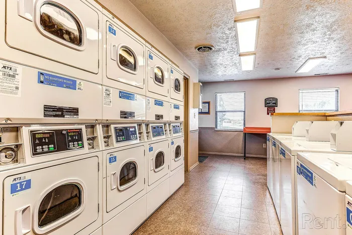 Modern apartment laundry room with multiple stacked washers and dryers, brightly lit with large windows and convenient folding area.