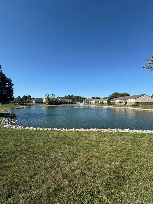 Serene community pond with a fountain surrounded by manicured lawns and modern apartment buildings under a clear blue sky.