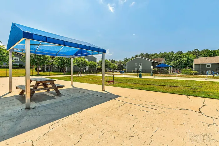 Inviting community outdoor space with picnic tables under a modern blue canopy, surrounded by lush greenery and cozy residential buildings.