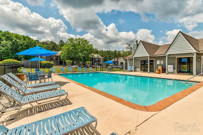 Inviting outdoor pool area with sun loungers, blue umbrellas, and charming clubhouse against a backdrop of lush greenery and blue skies.