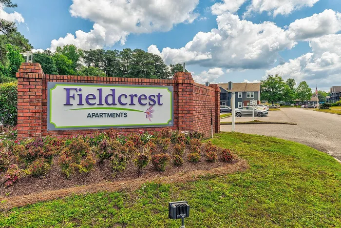 Inviting entrance of Fieldcrest Apartments with beautiful landscaping, a welcoming sign, and spacious parking under a bright, blue sky.