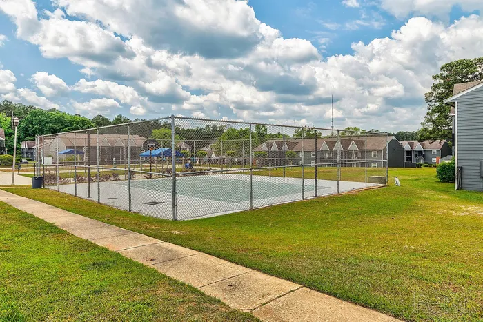 Inviting apartment community featuring a fenced tennis court surrounded by lush greenery and charming residential buildings under a bright, cloudy sky.