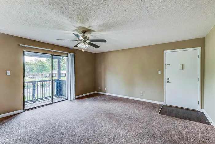 Bright and airy apartment living room with plush carpet, ceiling fan, and sliding door opening to a charming outdoor view.