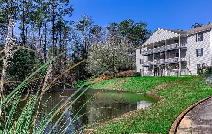 Serene lakeside apartment building nestled in lush greenery with expansive balconies and scenic views of tall trees under a clear blue sky.