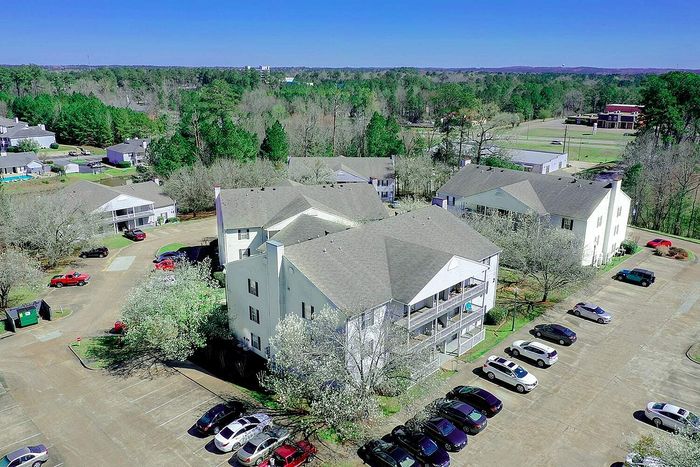 Aerial view of charming apartment complex nestled in a serene wooded area, featuring spacious balconies and ample parking space.
