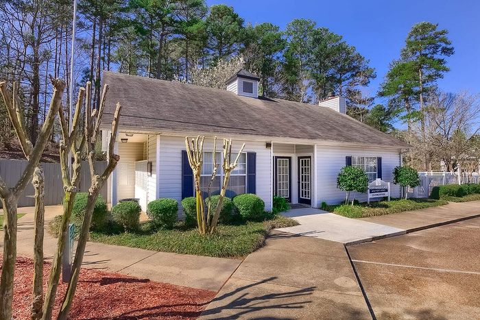 Charming leasing office with classic white siding, surrounded by lush greenery and tall trees, under a bright blue sky.