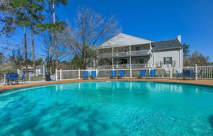 Inviting outdoor pool with sun loungers, surrounded by lush trees and charming white clapboard buildings under a clear blue sky.