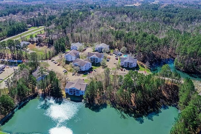 Scenic aerial view of charming apartment complex surrounded by lush greenery and tranquil lake, offering serene living in a natural setting.