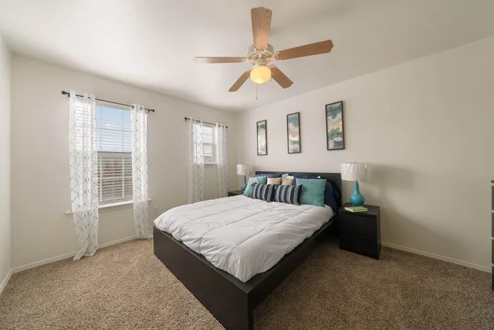 Bright and inviting bedroom with cozy carpet, modern ceiling fan, and elegant decor. Abundant natural light streams through large windows with sheer curtains.