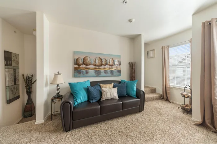 Inviting living room with plush carpeting, a cozy brown sofa adorned with vibrant blue pillows, and abundant natural light streaming through large windows.