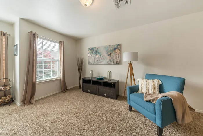 Bright and cozy living space with a soft carpet, blue accent chair, and elegant decor. Large window with curtains allows natural light to enhance the inviting atmosphere.