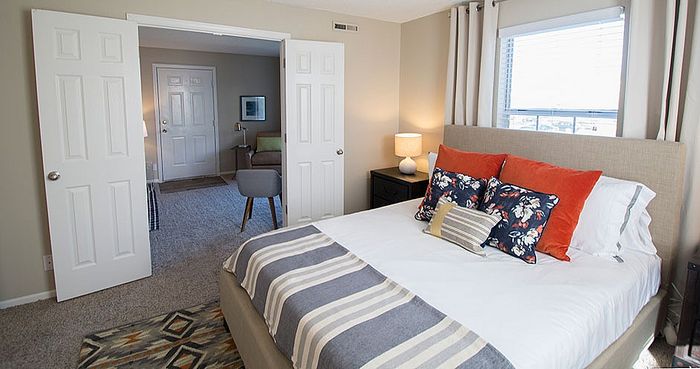 Cozy bedroom in modern apartment featuring a plush bed with vibrant pillows, large window with ample natural light, and access to a spacious sitting area.