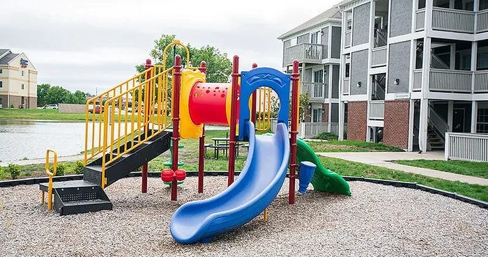 Inviting apartment complex featuring a vibrant playground with colorful slides surrounded by lush greenery and a scenic pond, ideal for family living.