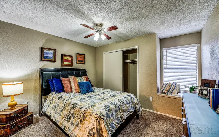 Cozy bedroom with soft lighting, modern ceiling fan, plush bedding, decorative artwork, and ample natural light from large window.