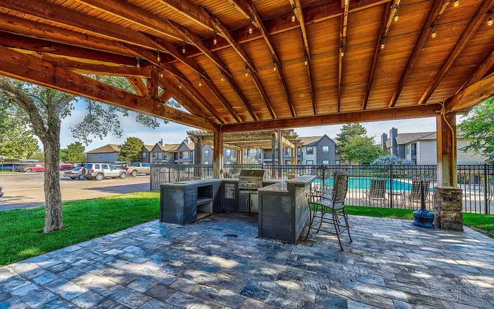 Charming outdoor living space with a wooden pergola and stylish stone patio overlooking a sparkling pool and apartment complex.