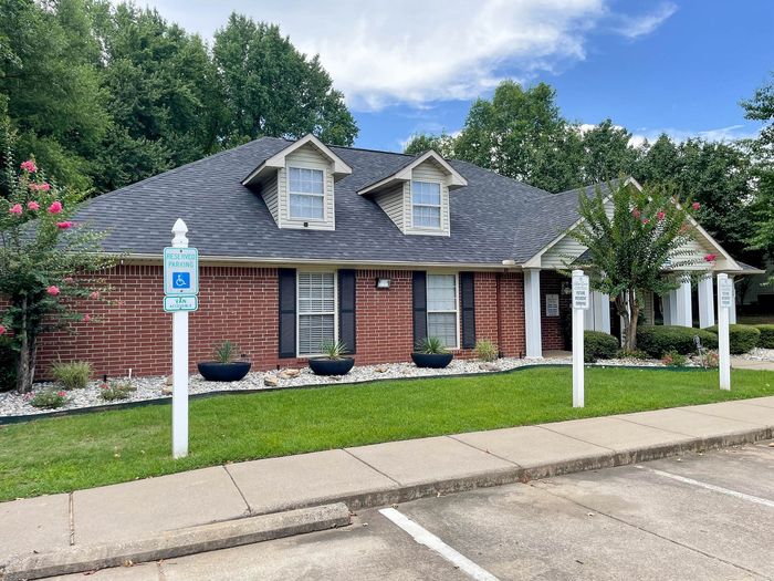 Charming brick apartment building with manicured lawn, accessible parking, and lush greenery against a bright blue sky.