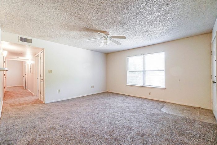 Bright and spacious living area with plush carpet, large window, and ceiling fan, perfect for comfort and relaxation.