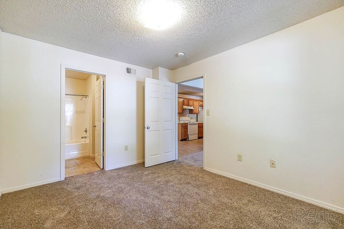 Inviting apartment interior showcasing a cozy carpeted bedroom with an ensuite bathroom and a glimpse of the spacious, light-filled kitchen.