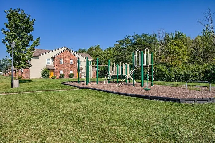 Charming residential community with an inviting playground surrounded by lush greenery and a picturesque brick and siding apartment building.