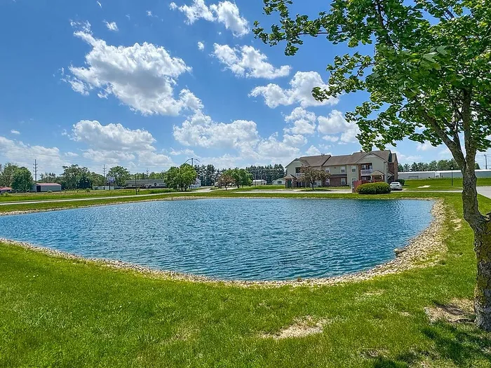 Scenic lake view from apartment community, featuring lush greenery, a tranquil pond, and a charming residential building under a bright blue sky.