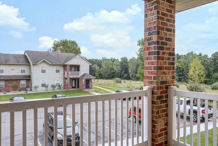 Charming residential view from balcony showcasing a peaceful suburban setting with a blend of brick and siding architecture, surrounded by lush greenery and ample parking space.