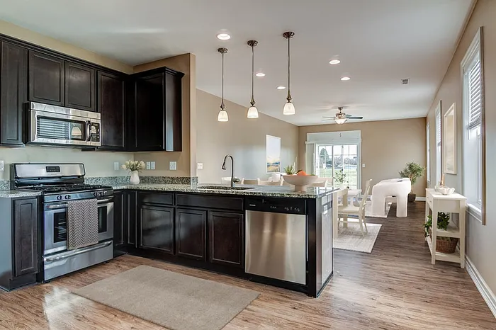 Modern open-concept kitchen and living area with sleek dark wood cabinetry, stainless steel appliances, and pendant lighting, leading to a bright dining space with large windows.