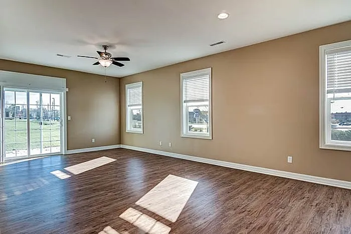 Bright and spacious living room with elegant hardwood floors, ceiling fan, and abundant natural light from large windows and sliding glass doors overlooking a scenic view.