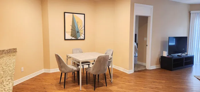 Cozy apartment dining area featuring a modern art piece, sleek table with four upholstered chairs, and warm-toned laminate flooring leading to a bright living room with natural light.