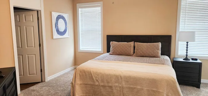Cozy bedroom with plush bedding, soft carpet, elegant artwork, and warm natural light filtering through the windows.