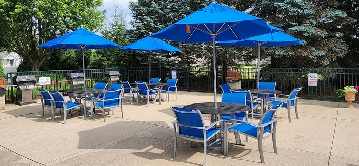 Inviting outdoor patio area with vibrant blue umbrellas and modern seating, perfect for social gatherings and relaxation. Surrounded by lush greenery, this space features multiple BBQ grills for an enhanced outdoor living experience.