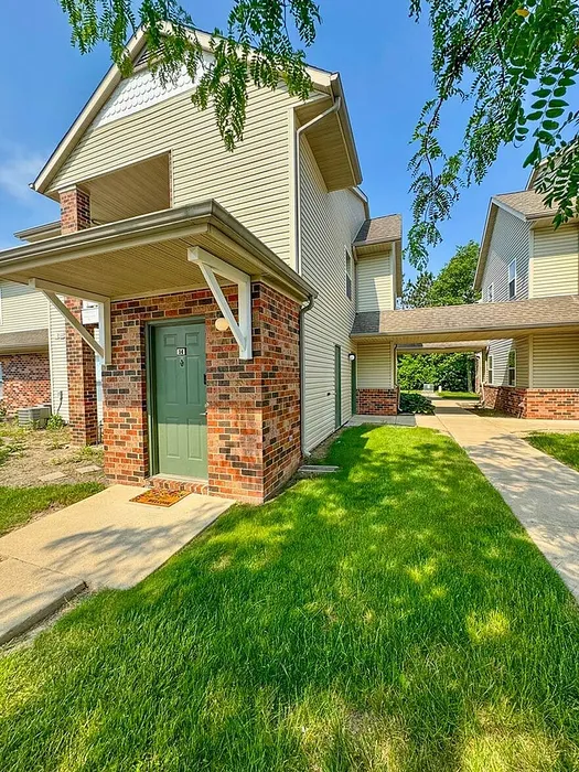 Charming two-story townhouse with brick accents and a welcoming green door, surrounded by lush greenery and sunny skies.