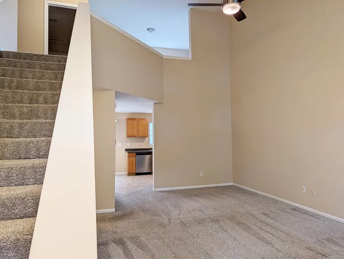 Inviting open-concept living area with plush carpet, modern ceiling fan, and a cozy staircase leading to an upper level. Adjacent kitchen features sleek wooden cabinetry and stainless steel appliances.