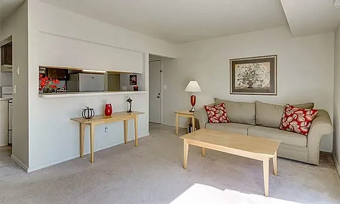 Bright and inviting living room with a cozy beige sofa, decorative cushions, and a light wood coffee table, adjacent to a partially open kitchen.