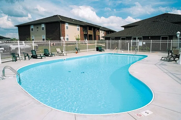 Inviting outdoor pool area with sparkling blue water, surrounded by a secure white fence and modern apartment buildings in the background, offering a perfect relaxation spot.