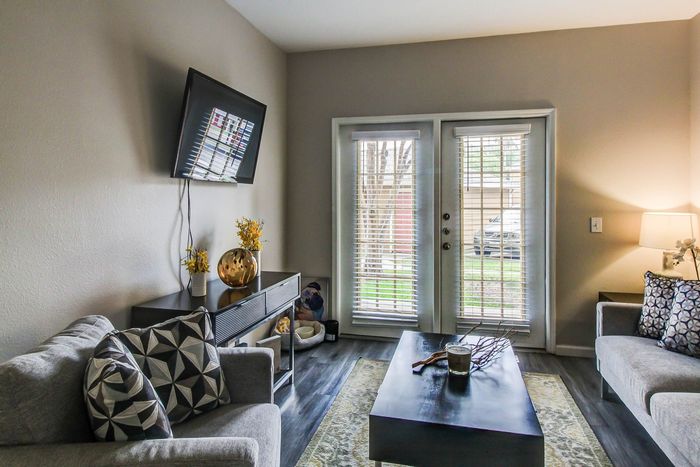 Cozy and modern living room with stylish decor, featuring a wall-mounted TV, plush sofas, and a sleek coffee table. Natural light streams through elegant French doors with blinds, creating a warm and inviting atmosphere.