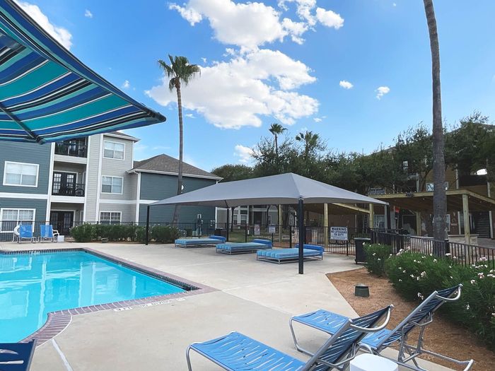 Inviting apartment complex with sparkling pool, stylish lounge chairs, and tropical palm trees under a bright blue sky.
