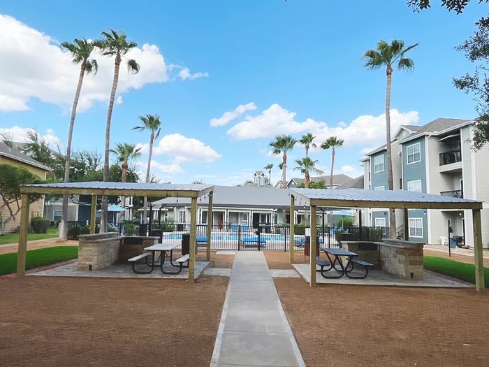 Inviting outdoor communal area with shaded seating, picturesque palm trees, and a refreshing pool surrounded by modern apartment buildings under a bright blue sky.