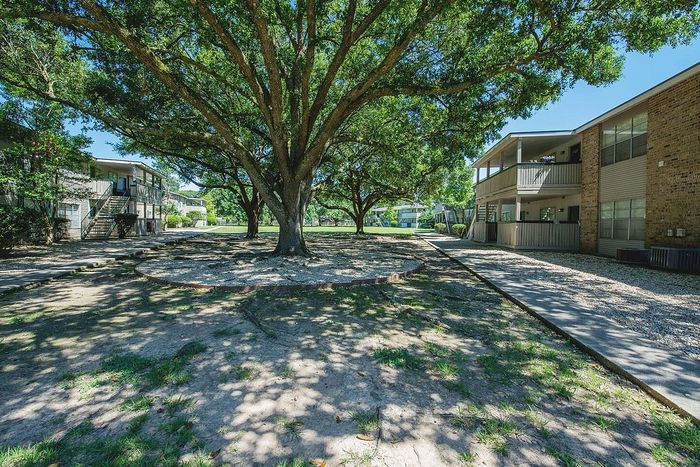 Charming apartment complex with lush greenery, featuring large, mature trees and well-maintained pathways perfect for a serene living environment.